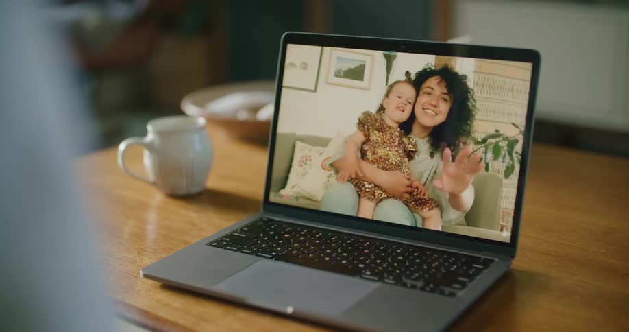 Person makes a video call on laptop with a woman with curly hair and holding a toddler girl in the living room at home