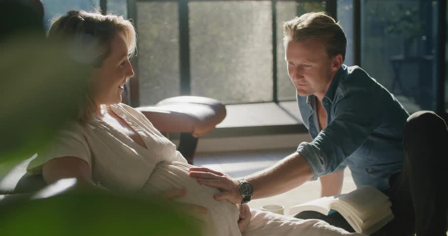 A man rests his hand on the stomach of a pregnant woman as they sit together on the floor of a house during the day