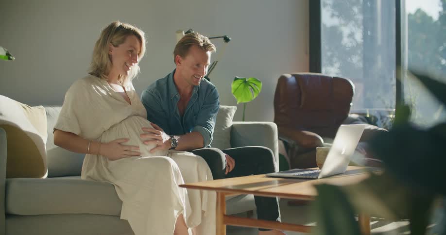 Young pregnant couple sitting in the living room talking on a video call on a laptop