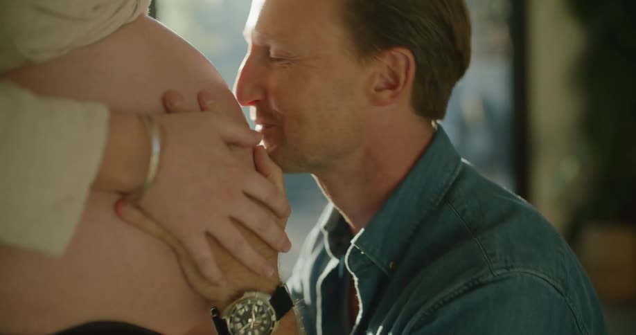 A man and a woman in a room as the man kisses the woman on her pregnant belly