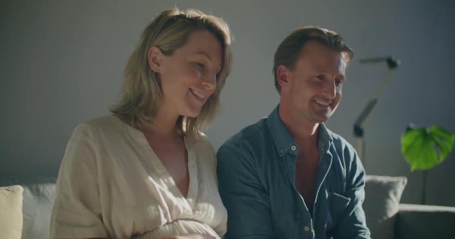 Young pregnant couple sitting in a couch in the livingroom greeting people on a video call