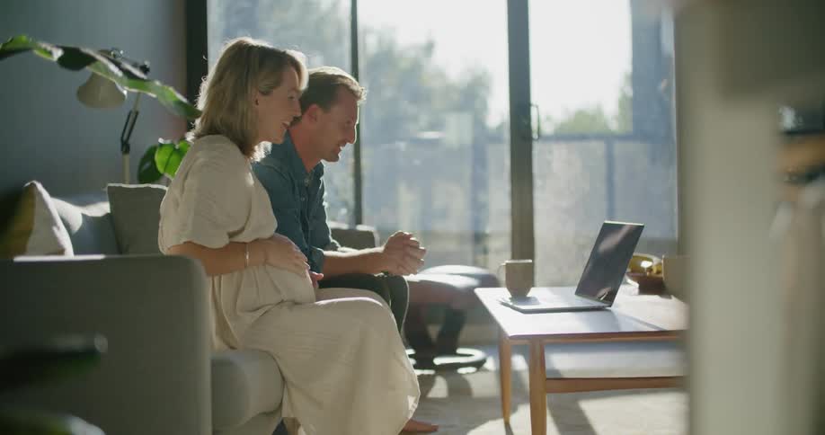 A man puts his coffee cup down so he can join his wife in waving at the person they are meeting with on a laptop screen