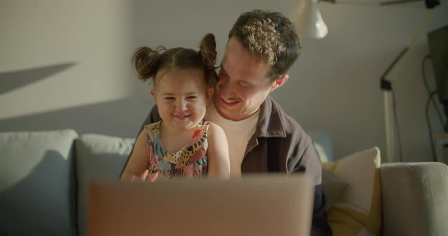 A man and toddler girl sit on a couch and happily wave to a laptop