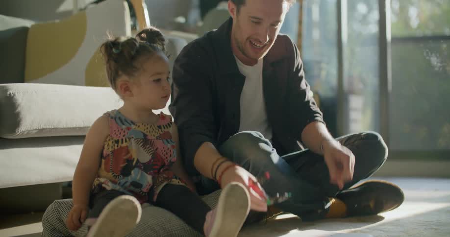 A man prepares a marker and sheet for paper for a young girl sitting beside him on a cushion on the floor of the living room of a house during the day