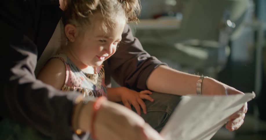 A holding a small girl between his arms and legs shows her a sheet of paper that causes her to laugh
