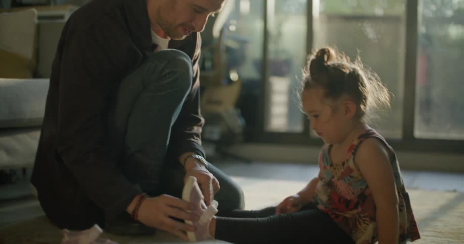 A man kneeling next to a young girl helps her to put on her shoes in the living room of a house