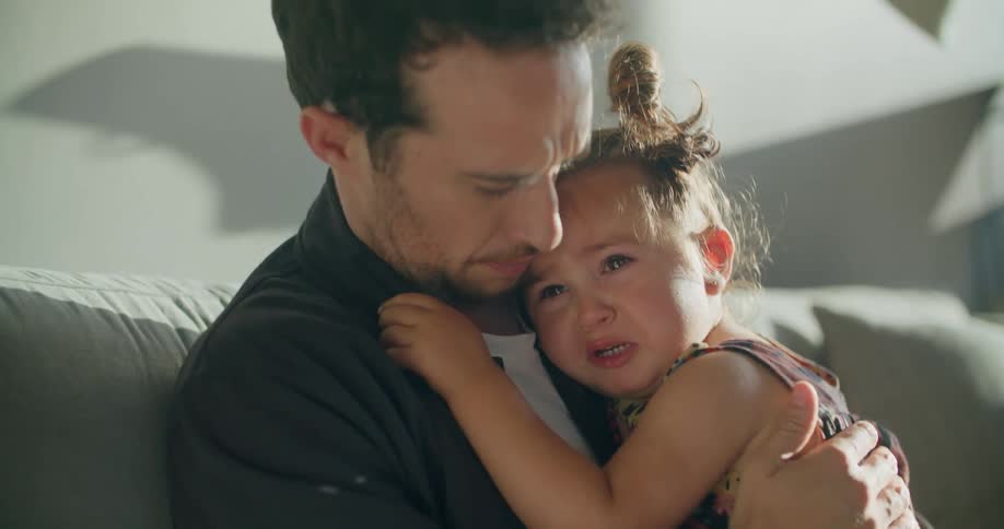 A man in a room hugging a girl who is crying with a couch a lamp and a wall in the background