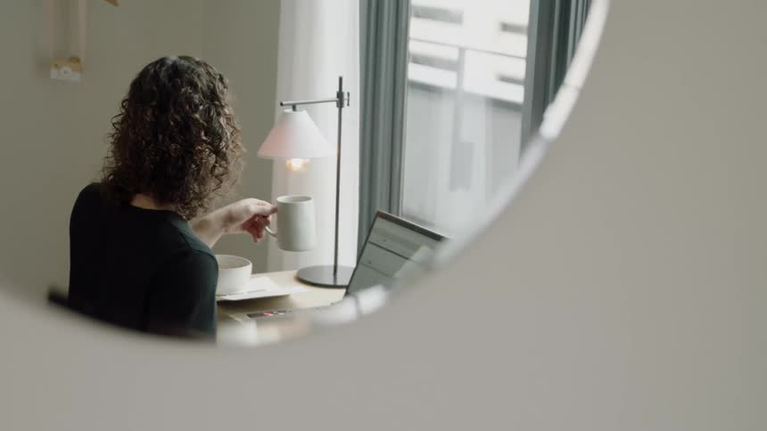 Person drinking coffee and sitting in front of laptop is reflected in mirror