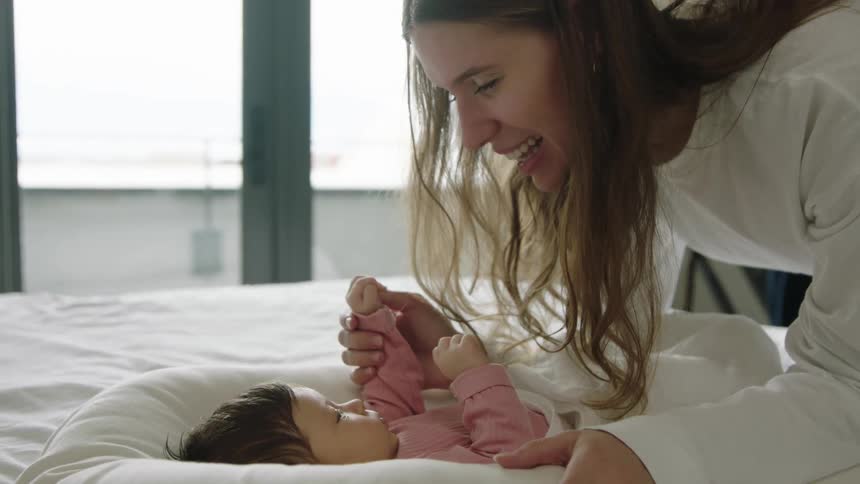 Mother laughs while playing with her infant son in the bed at home