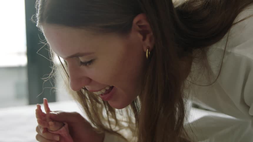 A woman kisses the fingers of a baby lying on a bed in bedroom during the day