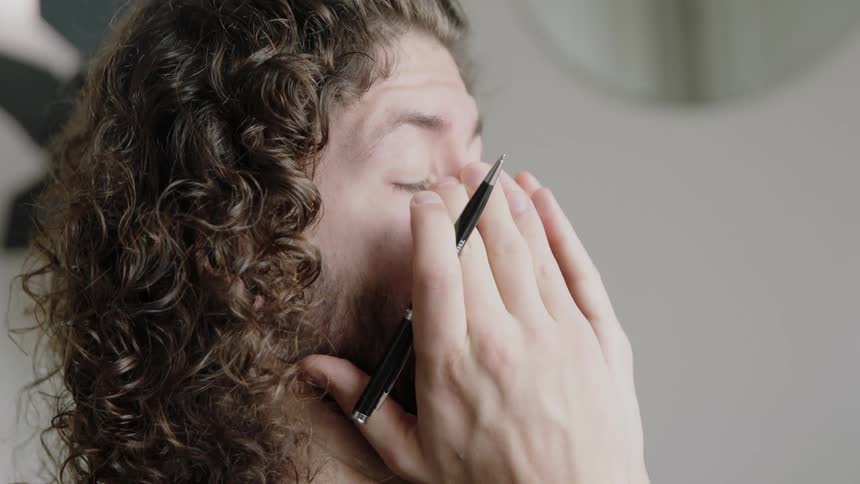 A man in a room holding a pen and rubbing his face with a wall and a mirror in the background