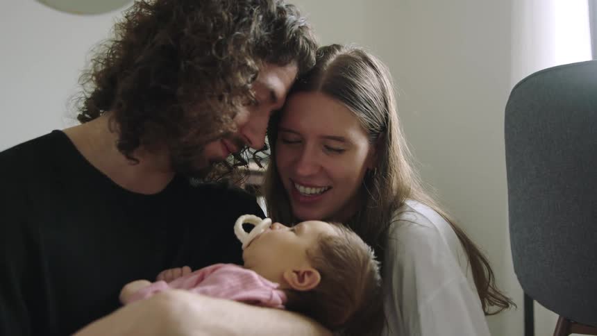 A man and woman sitting together indoors gaze at a baby as the man rocks her in his arms