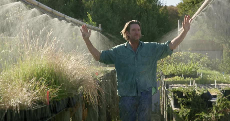 A man walks past the garden sprinklers reaching his arms out