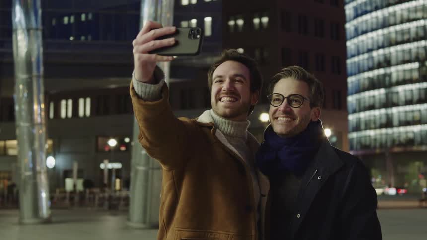 Two men stand close and smile as they take a selfie in a city at night