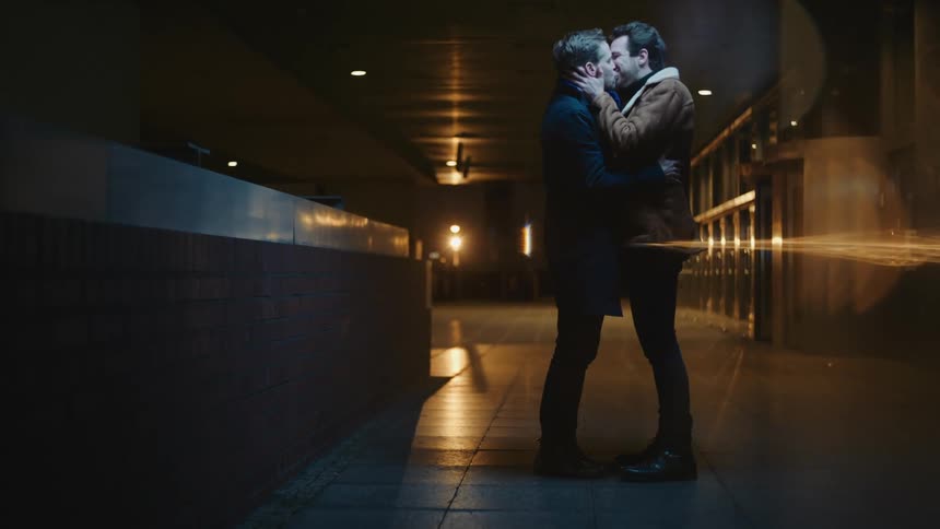 Two men stand in an empty concourse hugging and kissing