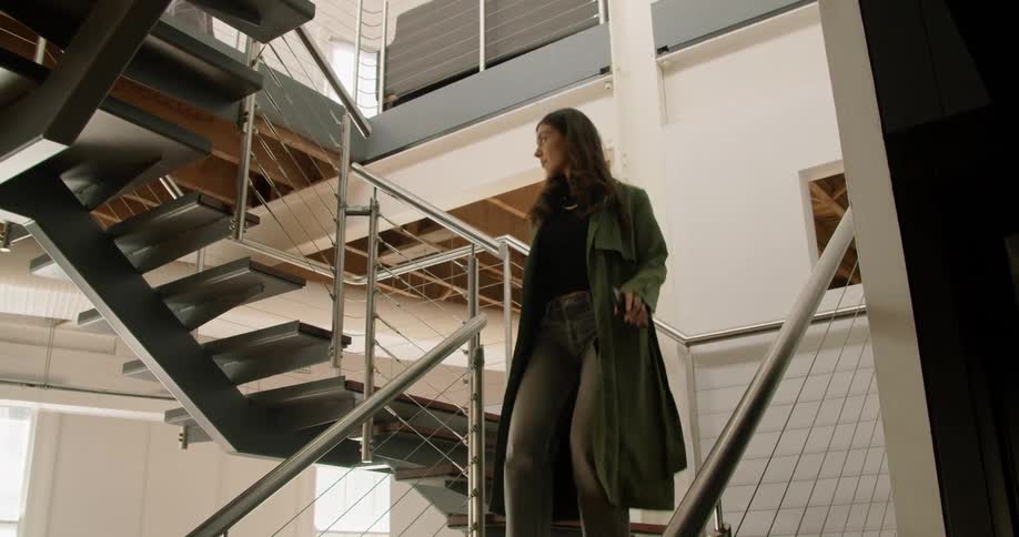 A woman checks her phone while walking down an open staircase with her raincoat on