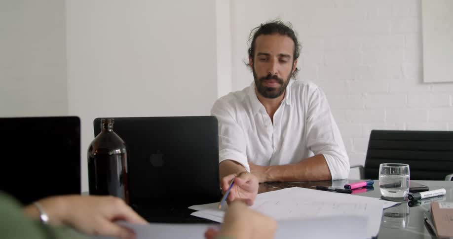 A man in a room holding a pencil sitting at a desk with a laptop talking to a woman