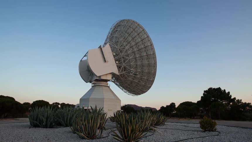 A satellite dish with plants and gravel in the foreground and trees the sky and clouds in the background