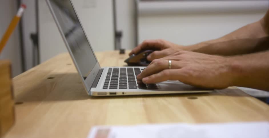 A man types on his laptop on a wooden desk