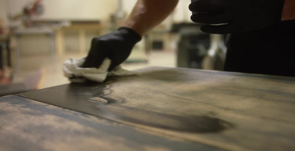 A man uses a rag to apply a dark stain on a long wood table
