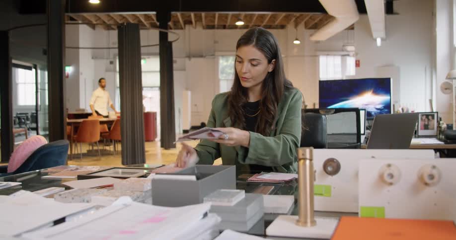 A woman sitting at a desk looking at fabrics and typing on a laptop with a wall and window in the background