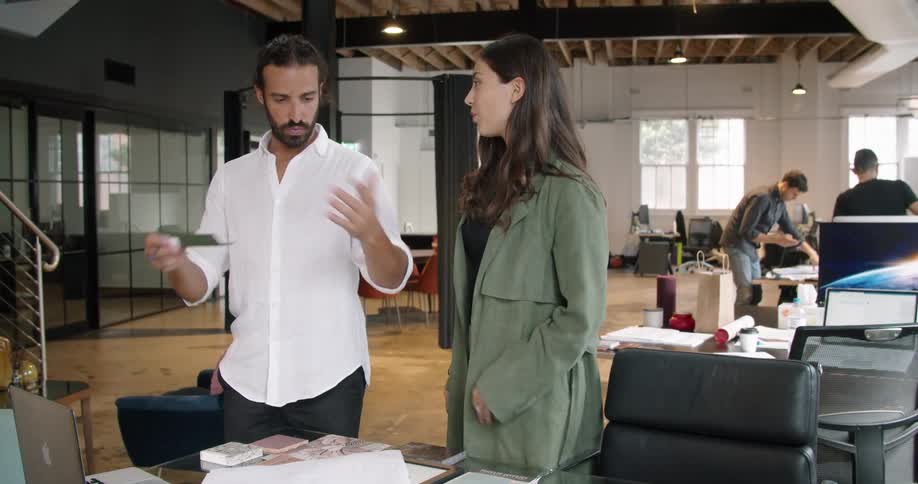 People walk around an office building a a man stand at a desk and shows a woman materials