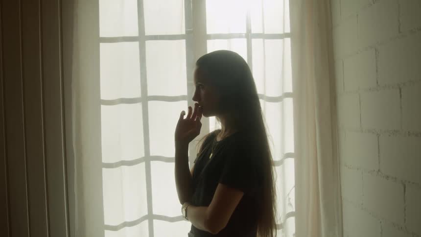 Woman with long brown hair and a black t shirt is silhouetted against the window