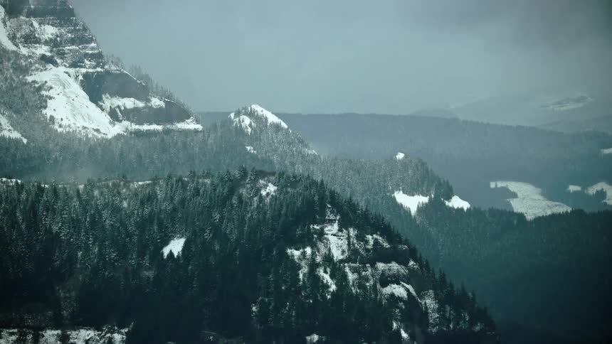 Clear cut logging is seen in the background of steep tree covered slopes in this airplane aerial shot in winter