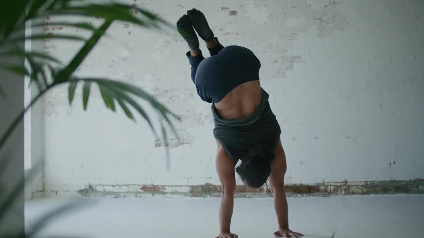 A man stretching and doing a handstand with a plant in the foreground and a wall in the background