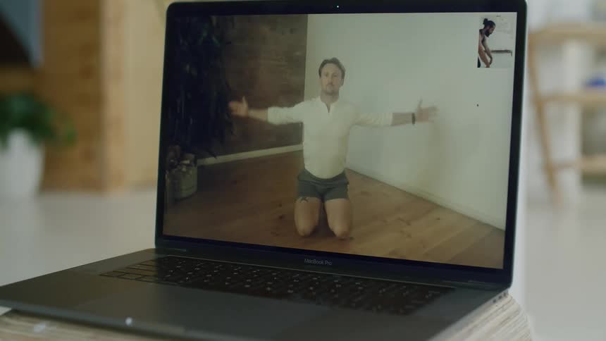 A man is displayed on a laptop screen kneeling on a floor stretching and exercising during a virtual class