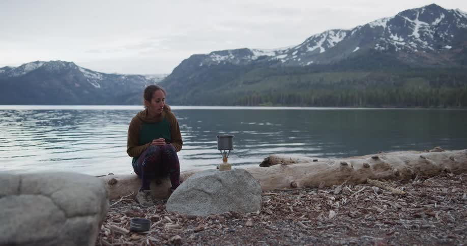 A woman sitting on a log by a lake with a mountain range the sky and clouds in the background