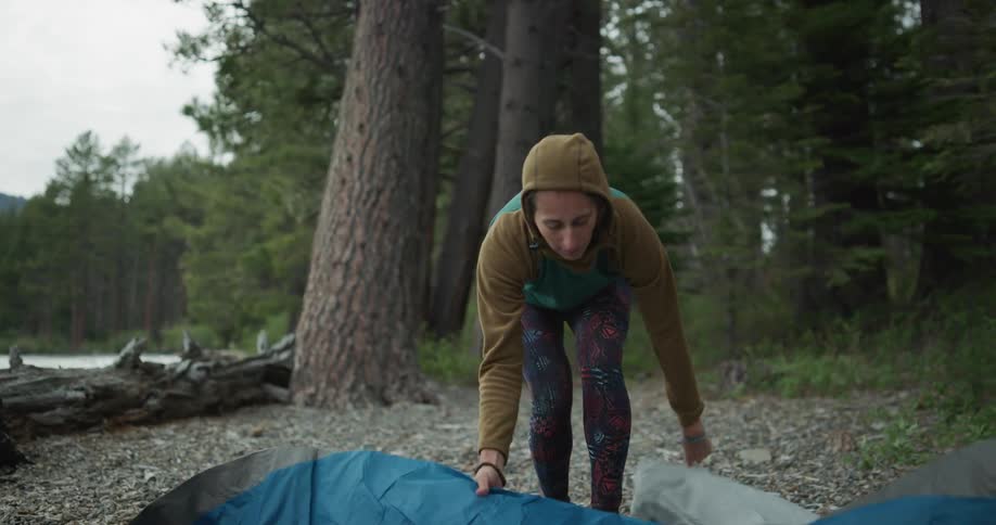 A woman in a campground next to a lake opening up a tarp with trees the sky and clouds in the background