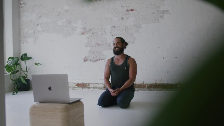 Man kneeling in a living room talking to a face cam on a laptop computer propped up on a wooden block