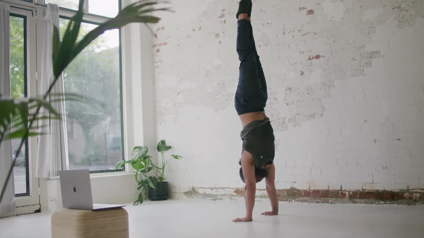 A man uses a laptop to participate in a video conference while he performs yoga poses and gives instruction