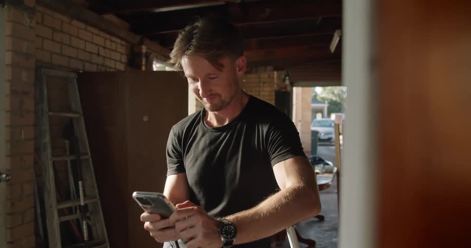 Blonde man with a black t shirt and a wristwatch uses a smartphone in the garage