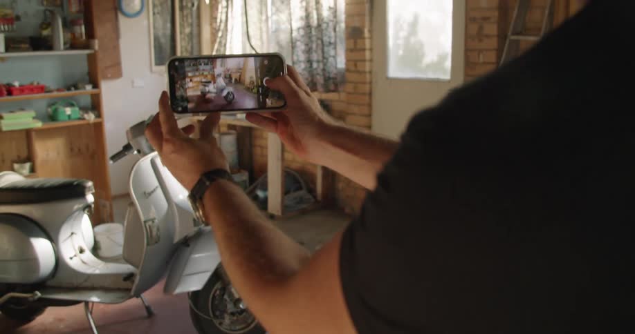 A man takes multiple photos of his silver scooter parked indoors with his smartphone