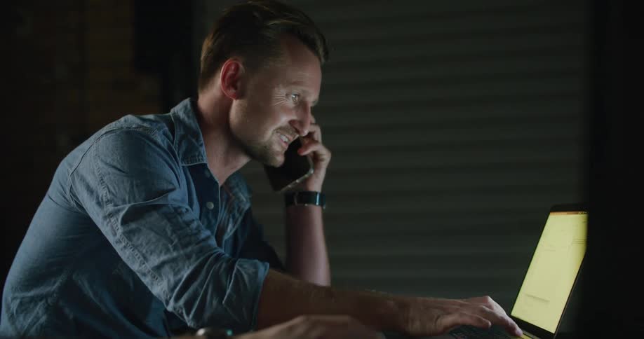 A man is talking on his cellphone as he uses a laptop