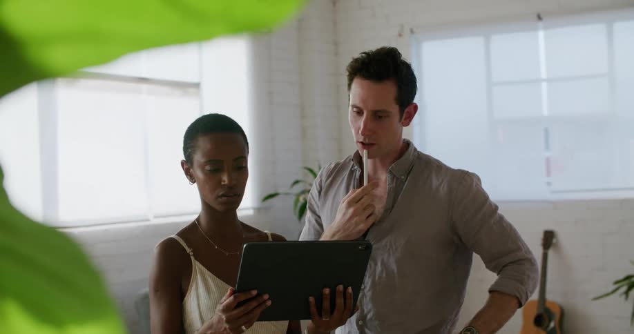 A woman holds a tablet in her hands while a man standing behind her looks over her shoulder at points at the screen indoors at daytime