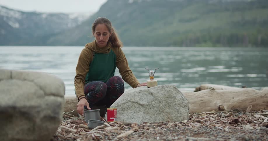 Woman sets up portable burner on shore across water from forest and mountains