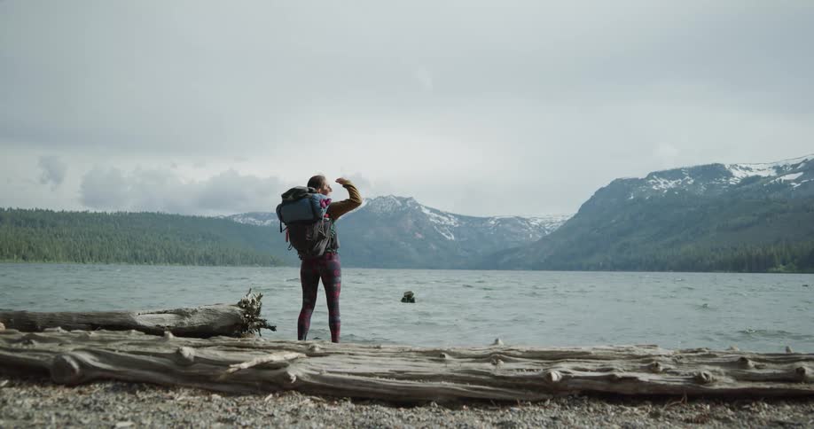 A woman with a backpack standing by a lake with a mountain range the sky and clouds in the background