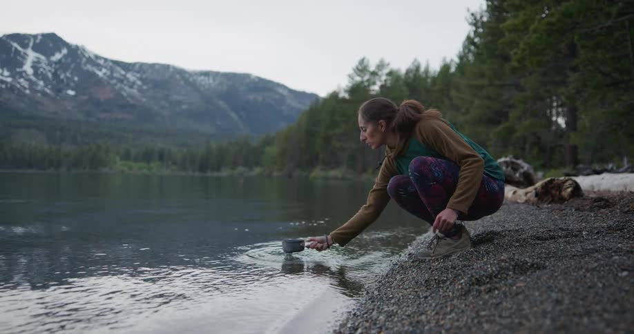 Woman scooping up water with metal cup at shoreline on a lake in the wilderness