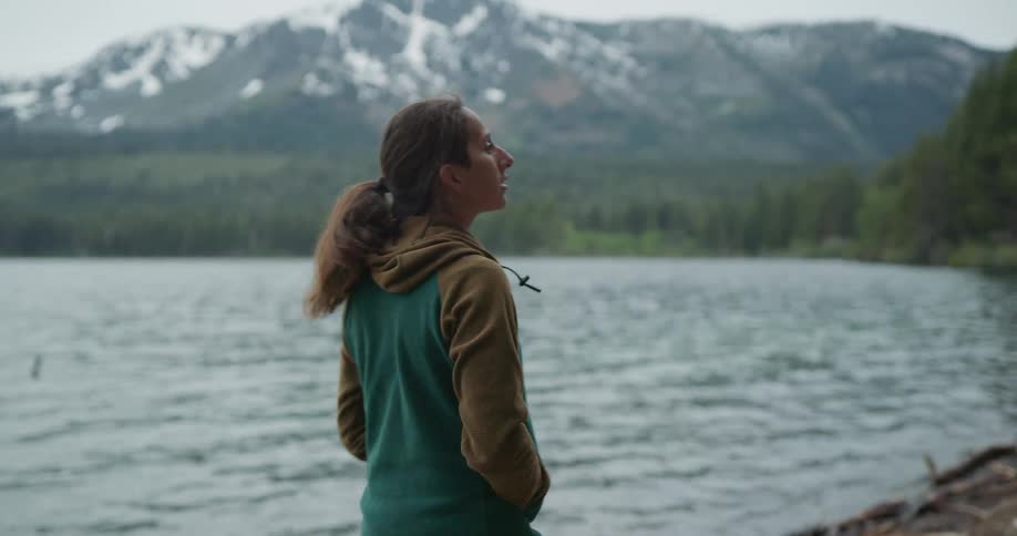 A young woman walks along the shore of a lake surrounding by forest during the day