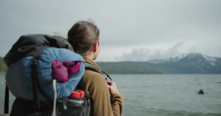 A backpacker smiles as she looks over a lake and toward a mountain range across the way