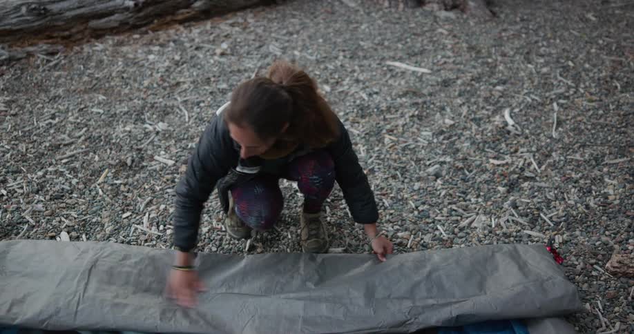A woman in a campground rolling up a tarp with a backpack and a log in the background