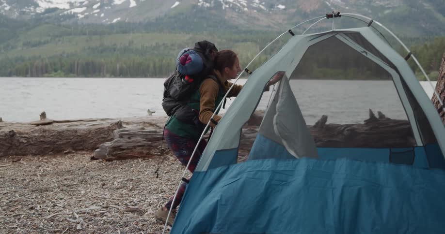 A woman with a backpack on walks to a tent on the shore and unzips it and takes her back pack off and puts it in the tent