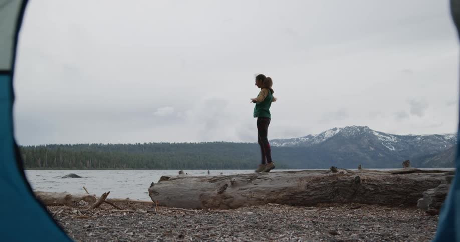 A woman tip toes across a tree trunk lying on the shore