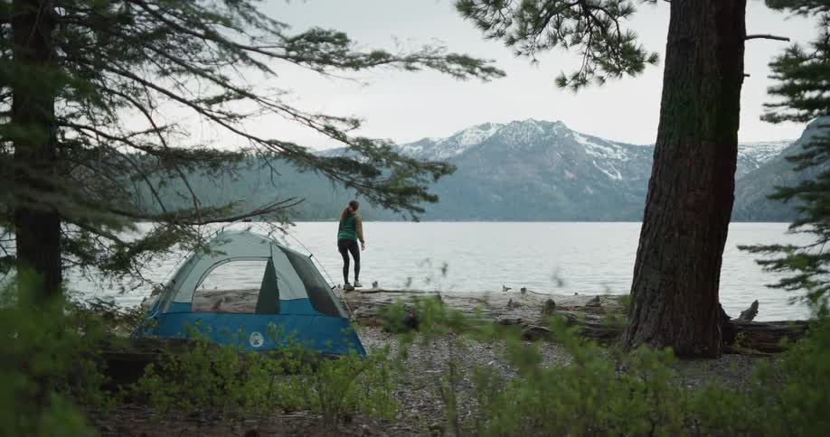 A backpacker stops near a camping site and walks to the edge of a lake to take in the view of a mountain