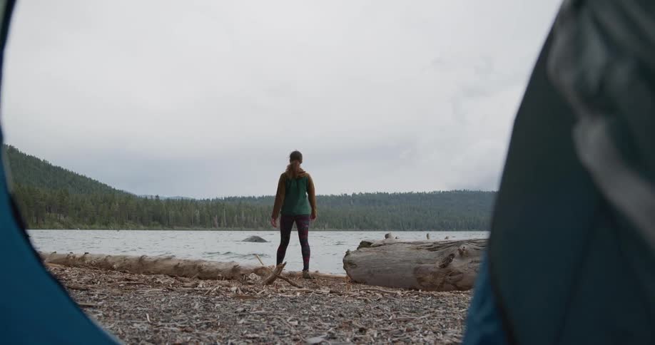 Woman opens tent and walks out to stretch while standing on shore across from forest