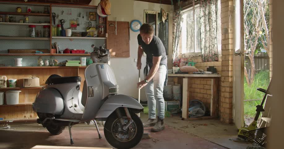 A man in a room sweeping the floor with a broom with a moped in the foreground and a wall and window in the background