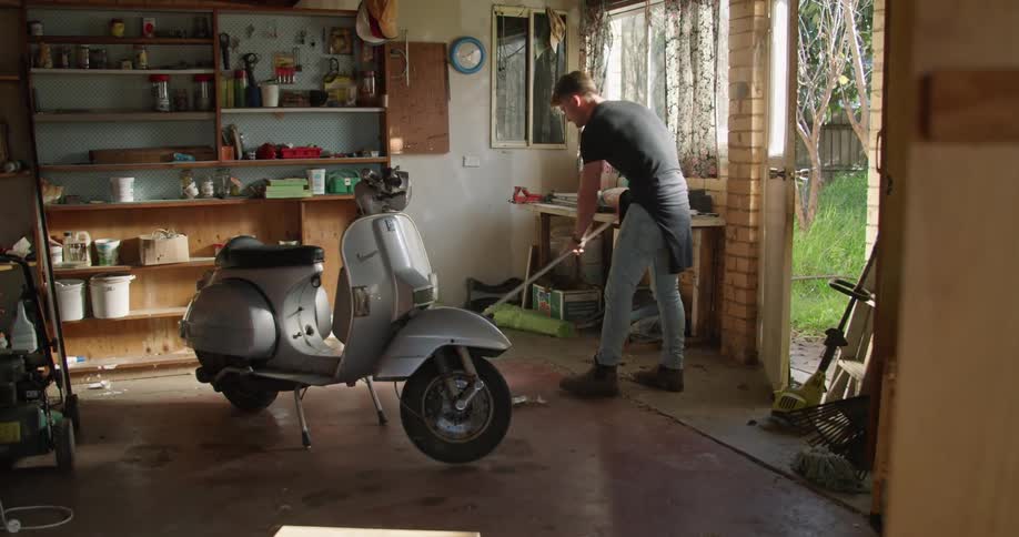 A man sweeping the garage floor with a broom around a vespa scooter